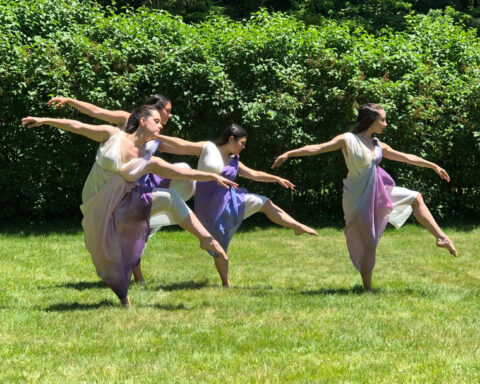 A photo of four women in flowing dresses, performing an elegant dance on a grassy lawn on a sunny day.