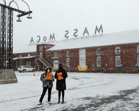 Mid-distance photo of two woman standing in a snowy parking lot holding bright orange flyers.