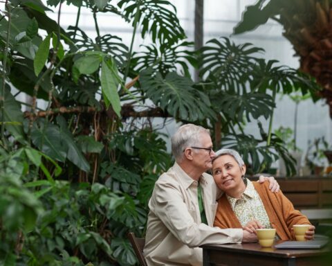 Elderly man and woman seated at a cafe-style table in an atrium or sunroom.