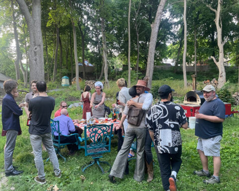 Exterior photo of a dozen or so people milling about during a picnic on a lawn under some trees.