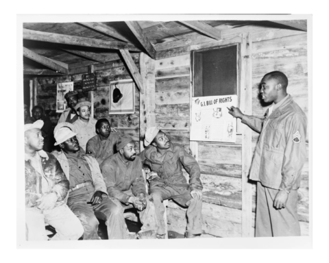 In a WWII barracks in Italy, a group of Black soldiers are seated as a fellow Black soldier explains a poster describing the G.I. Bill of rights.