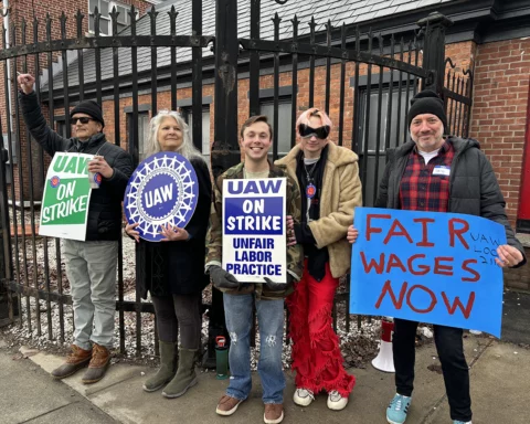 A diverse group of five MASS MoCA union members stand resolutely against a wrought-iron fence, each holding protest signs. From left to right: a man with a green 'UAW ON STRIKE' sign, a woman with a circular UAW sign, a young man with a 'UAW ON STRIKE UNFAIR LABOR PRACTICE' sign, another person in sunglasses and red fringed pants, and a man with a sign stating 'FAIR WAGES NOW.