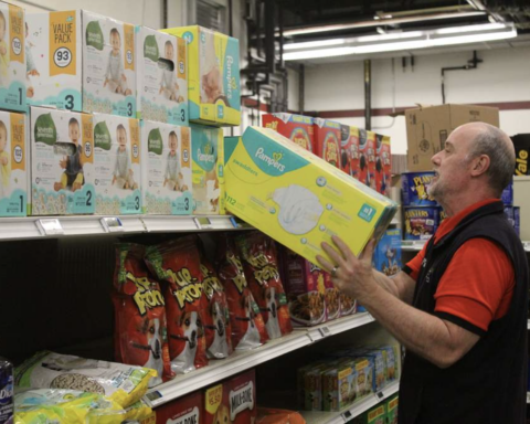 A male employee wearing a red vest over a black shirt is restocking a supermarket shelf with a box of Pampers diapers. The shelves are partially filled with various diaper packs and bags of dog treats. The lighting is artificial, and the environment suggests a warehouse-style store.