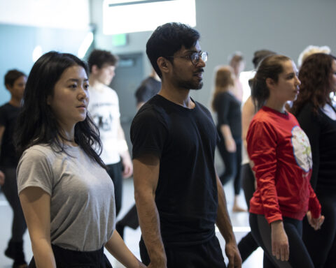 A diverse group of people practicing in a dance studio, focusing intently while standing in a line. The man in the foreground is wearing glasses and a black shirt, with the woman next to him in a gray t-shirt, both part of the workshop, "Tuning your Instrument.
