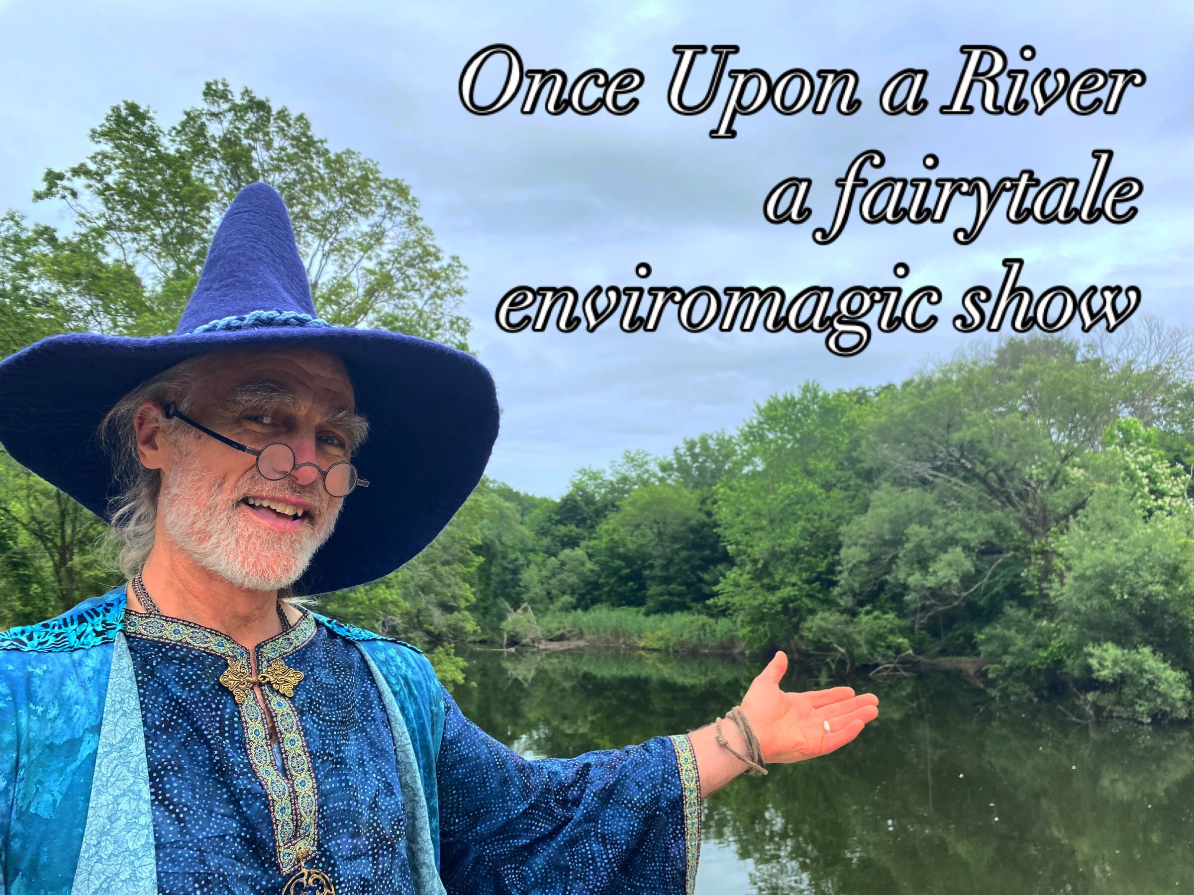 A smiling man in a blue wizard costume and hat stands beside a calm river, gesturing toward the water with one hand. Trees line the opposite bank, and overlaid text reads “Once Upon a River, a fairytale enviromagic show.”