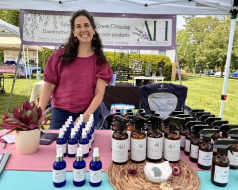 Monika of Aroma Home at a Lenox market event, with handmade home and body care products displayed under a vendor tent.